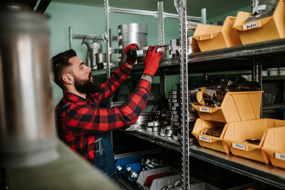Homme barbu en chemise à carreaux rouge et noir travaillant dans un entrepôt industriel, manipulant des pièces métalliques sur étagères modulaires